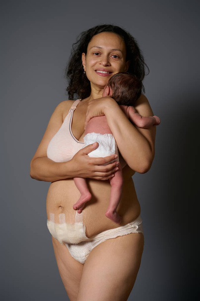 Ευτυχισμένη νέα μητέρα χαμογελά κοιτάζοντας κάμερα, κρατά το νεογέννητο μωρό της και δείχνει γυμνή κοιλιά με επίδεσμο κρύβει ουλές μετά την καισαρική τομή και μετά τον τοκετό ελαττώματα όπως ραγάδες, απομονωμένη σε γκρι φόντο - Φωτογραφία, εικόνα