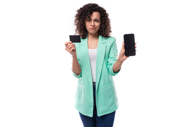 dobře upravená mladá brunetka kudrnatá obchodnice oblečená v modré bundě drží plastovou bankovní kartu mockup a smartphone. - Fotografie, Obrázek