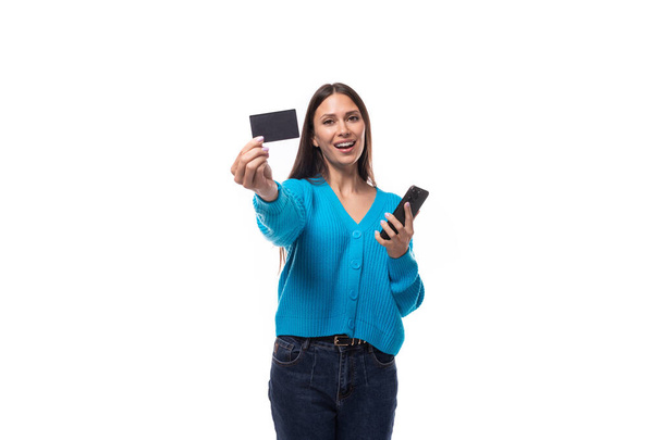 jong zelfverzekerd brunette vrouw met licht make-up dragen van een blauwe knop-down trui houdt een plastic kaart mockup en een smartphone in haar hand op een witte achtergrond. - Foto, afbeelding