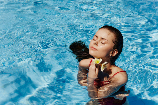 Молодая женщина в бассейне в красном купальнике с красивой улыбкой и балийским цветком на солнце плавает в бассейне, концепция релаксации на отдыхе. Высокое качество фото - Фото, изображение