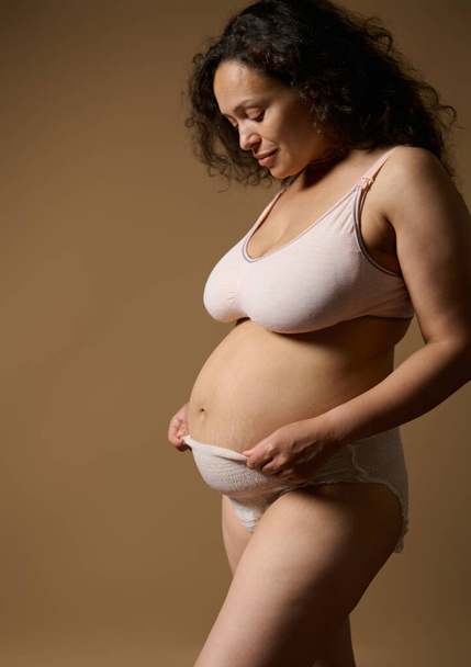Pewna siebie, piękna kobieta matka w bieliźnie, pozująca ze swoim pourodzeniowym nagim brzuchem z wadami i rozstępami kilka dni po porodzie, w odizolowanym beżowym studio. Odzysk poporodowy - Zdjęcie, obraz