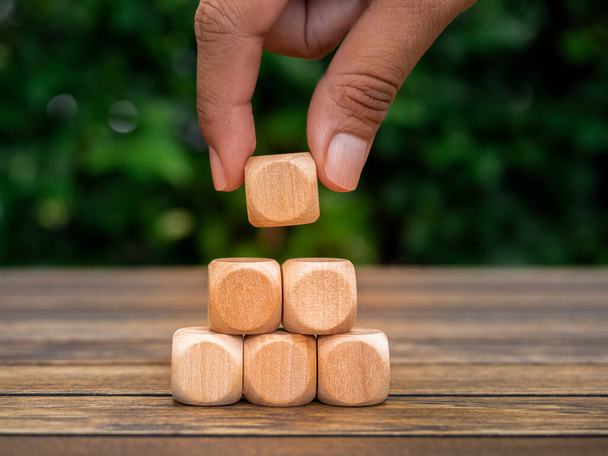 Empty blank wooden cube blocks arranged in pyramid shape, hand put the last one on top, on wood desk and green plant background. Business growth process, education, sustainable organizational concept. - Photo, Image