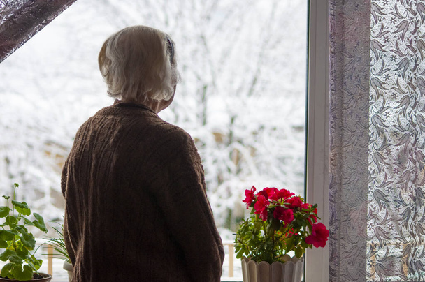Γριά μοναχική γυναίκα στέκεται κοντά στο παράθυρο του σπιτιού του και ψάχνει και ονειρεύεται το χειμώνα - Φωτογραφία, εικόνα