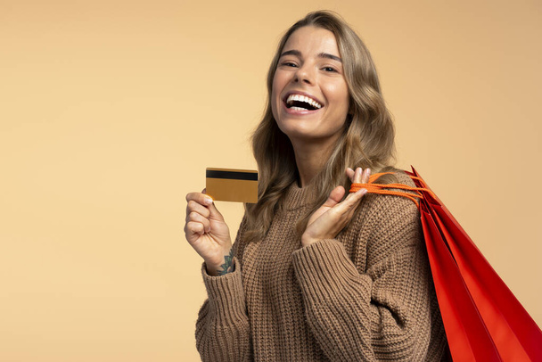 Retrato de mulher branca bonita sorridente segurando cartão de crédito e sacos de compras vermelhos isolados em fundo bege, espaço de cópia. Compras, venda, conceito de sexta-feira negra  - Foto, Imagem