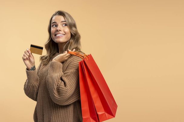 Piękna uśmiechnięta kobieta trzymająca kartę kredytową i czerwone torby na zakupy odwracając wzrok na beżowym tle, przestrzeń na ksero. Zakupy, sprzedaż, czarny piątek, koncepcja reklamy  - Zdjęcie, obraz