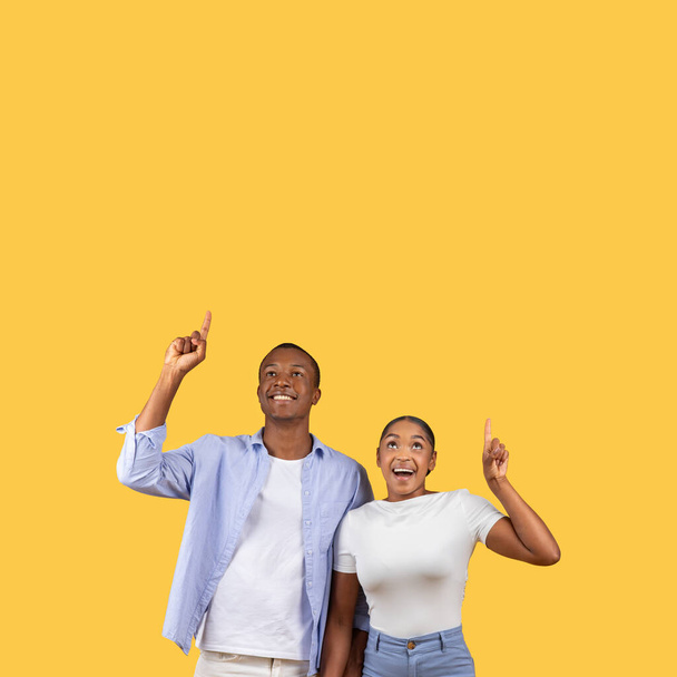 Emocionada joven pareja afroamericana ambos apuntando hacia arriba con la mirada de sorpresa y alegría en sus rostros, de pie sobre un fondo amarillo llano - Foto, imagen