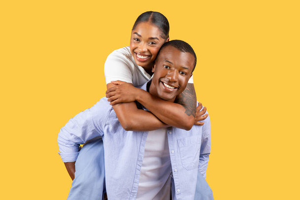 Зачарована чорна пара насолоджується їздою на поросяті, жінка сміється і обіймає чоловіка ззаду, встановлений на яскраво-жовтому тлі - Фото, зображення