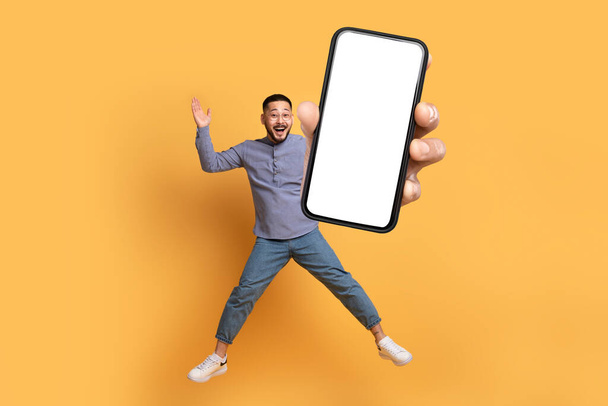 Смешной корейский парень прыгает, демонстрируя огромный телефон с пустым сенсорным экраном, идеально подходит для современного интернета и бизнес-концепций, студия снята на ярком желтом фоне. Полная длина - Фото, изображение