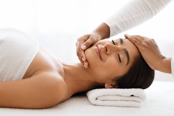 Una serena dama india sonriente recibe un masaje facial calmante, enfocándose en su frente y barbilla, de un terapeuta experto en masajes en un ambiente de spa lujoso, primer plano - Foto, imagen