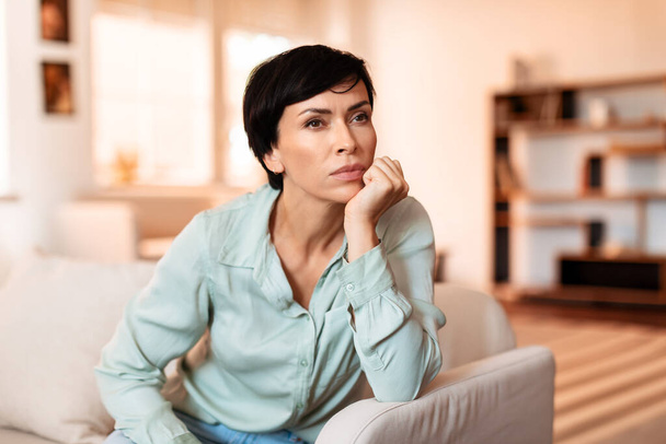 Middle aged woman sitting thoughtfully on her living room sofa at home, posing with a serious look, her pensive expression reflecting deep concern. Moment of reflection and introspection - Photo, Image