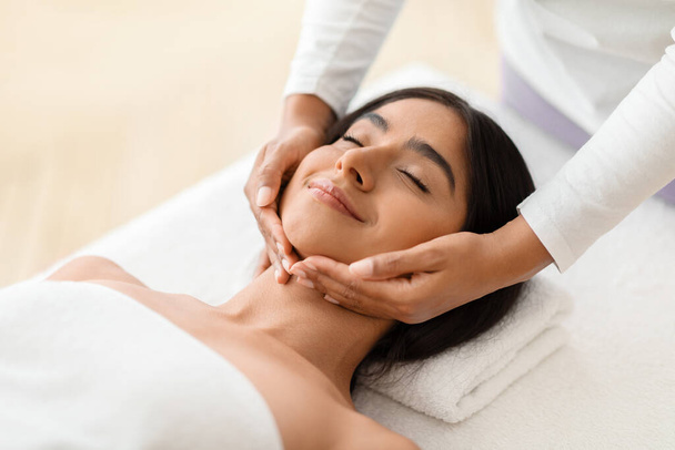 Beautiful young indian woman enjoying face lifting massage in spa salon, professional therapist making skin lift procedure to relaxed brunette lady lying on table in wellness center, closeup - Photo, Image