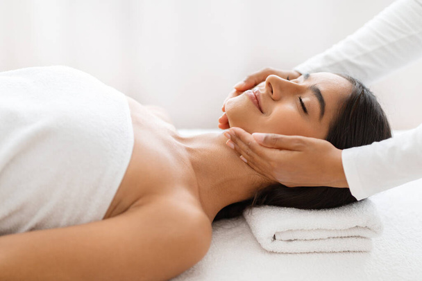 Relaxed pretty young indian woman enjoying face lifting massage in spa salon, therapist hands making skin lift procedure to relaxed brunette lady lying on table in wellness center, side view - Photo, Image
