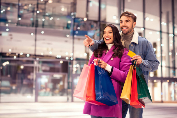 Wesoła para kupujących z kartą kredytową gotowa na świąteczne zimowe zakupy przed nowoczesnym sklepem handlowym, wskazując na noworoczną ofertę sprzedaży i wolną przestrzeń, na tle miejskiego wieczoru - Zdjęcie, obraz