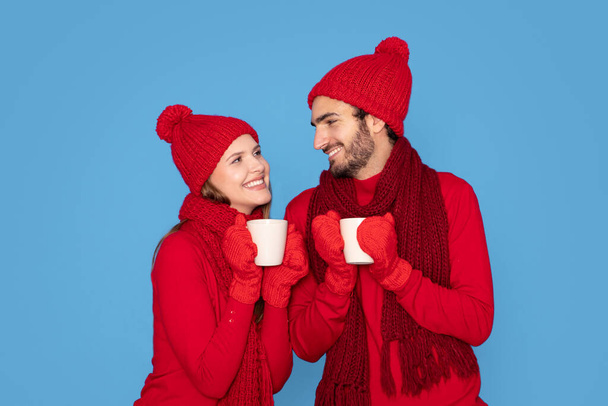 Joven pareja romántica usando manoplas de punto y sombreros bebiendo cacao juntos, hombre feliz y mujer sosteniendo tazas con chocolate caliente y sonriendo el uno al otro, posando aislados sobre fondo azul - Foto, imagen