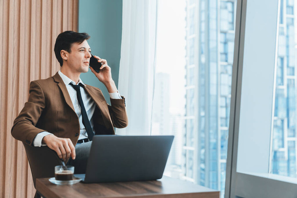Κοντινό πλάνο ενός όμορφου επιχειρηματία που τηλεφωνεί στον διευθυντή ενώ κάθεται κοντά στο παράθυρο με θέα στον ουρανοξύστη. Εκτελεστικός διευθυντής μιλάει εργασίας με τη χρήση τηλεφώνου και laptop. Κοίτα στην άκρη. Διακοσμημένα. - Φωτογραφία, εικόνα