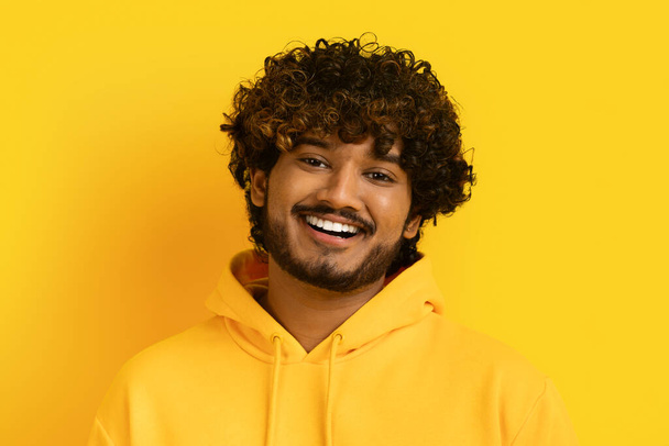 Closeup studio shot of positive handsome curly bearded young indian guy wearing yellow hoodie smiling at camera isolated on yellow background. Human emotions concept - Foto, Bild