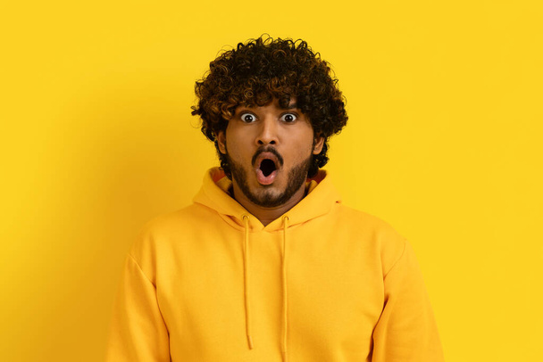 Portrait of shocked bearded curly young indian guy with mouth and eyes wide open wearing yellow hoodie posing isolated on yellow studio background, expressing strong emotions - Foto, immagini