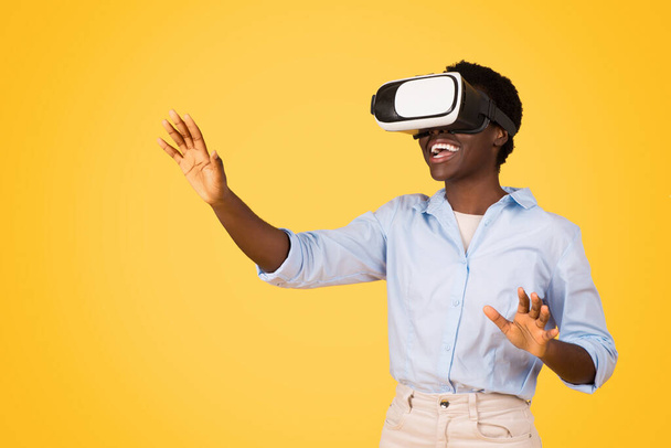 Sonriente joven afroamericana estudiante americana en gafas casuales y vr, controla el espacio de copia con las manos, disfrutar del juego, aislado en el fondo del estudio amarillo. Estudio y educación, realidad virtual - Foto, Imagen