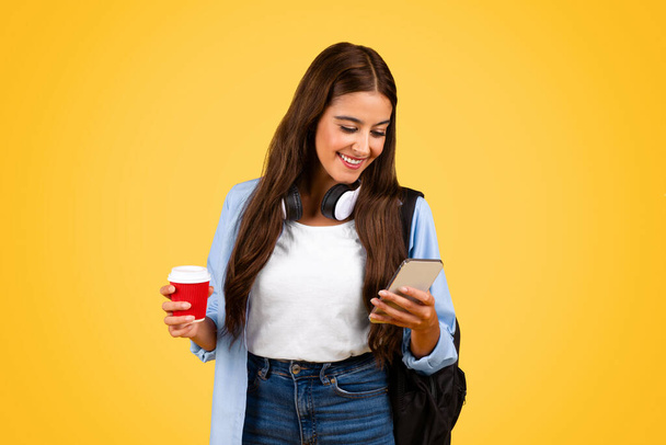 Happy teenager student woman with mobile phone, chatting, enjoy cup of coffee takeaway, isolated on yellow background. Spare time with online app, study break and social networks, music - Фото, изображение