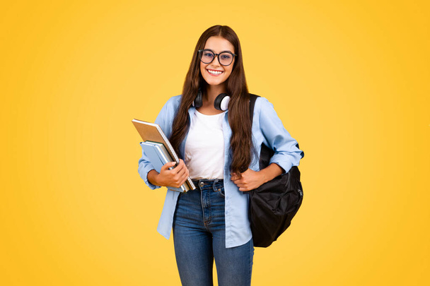 Positive nerd teenager caucasian student lady in glasses, with backpack, books enjoy study, isolating on yellow studio background. Pupil lifestyle at school, exam, education - Photo, Image