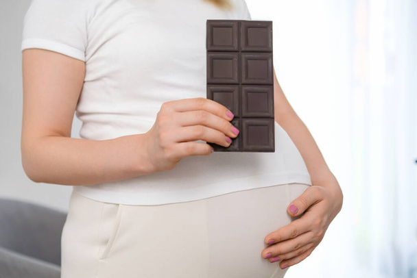 Una mujer embarazada sostiene una barra de chocolate y acuna su estómago. Dulces durante el concepto de embarazo - Foto, imagen