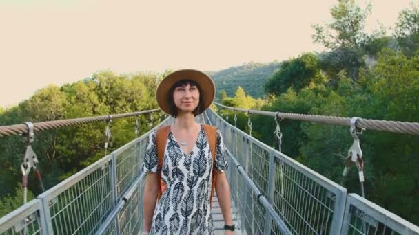 Dívka v klobouku a s batohem kráčí po mostě přes řeku. Mladá žena kráčí po visutém mostě přes řeku před podzimním lesem. Holka v klobouku. cestování a dobrodružství. - Záběry, video