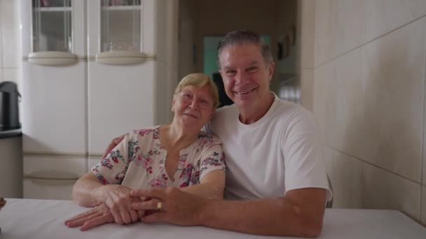 Boldog idős anya pózol kamerának a felnőtt középkorú fia mellett otthon a konyhában. Valódi emberek a családi életmód jelenetben - Felvétel, videó