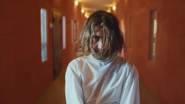 Mężczyzna z brudnymi włosami cierpiący na zaburzenia psychiczne noszący kaftan bezpieczeństwa stojący w szpitalnym korytarzu patrząc przed kamerą - Materiał filmowy, wideo