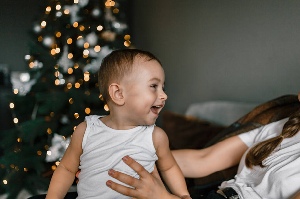 Un primo piano di un bambino felice sullo sfondo di un albero di Natale. Il bambino è a casa in un'atmosfera festiva accogliente. Vacanze invernali e concetto di persone - Foto, immagini