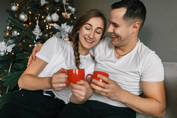 A young and happy couple sits near the Christmas tree at home. The beautiful man and woman enjoy hot chocolate and spend the New Year holidays in a comfortable home atmosphere - Photo, Image