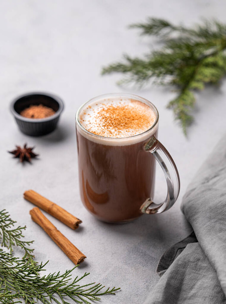 Γυάλινη κούπα με ζεστή σοκολάτα και αφρό γάλακτος σε ελαφρύ φόντο με ξυλάκια κανέλας, γλυκάνισο και κλαδιά πεύκου. Θερμαινόμενο χειμώνα Χριστουγεννιάτικο ποτό έννοια. - Φωτογραφία, εικόνα