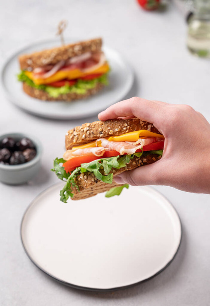 Uma mão segura um sanduíche torrado de pão de grão com queijo cheddar e bacon recheado com tomate e alface em um fundo leve com ingredientes de perto. Conceito de pequeno-almoço saudável e dietético.  - Foto, Imagem
