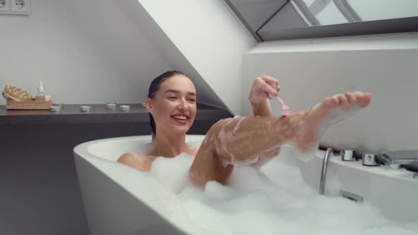 Boeiende 4K slow-motion video toont een vrolijke vrouw ondergedompeld in een schuimig bad, scheren haar benen. De beelden belichten de mix van ontspanning en persoonlijke verzorging in een serene omgeving - Video