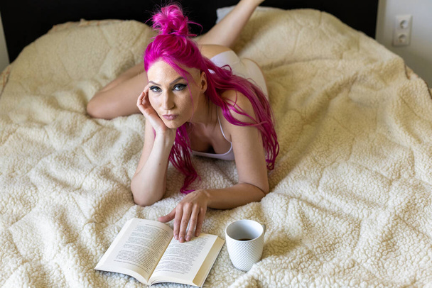 Γοητευτική νεαρή γυναίκα με σέξι εσώρουχα και ροζ μαλλιά ξαπλωμένη στο κρεβάτι με βιβλίο και φλιτζάνι καφέ - Φωτογραφία, εικόνα