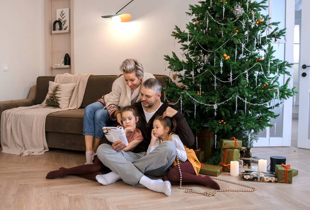 Πορτρέτο μιας φιλικής οικογένειας διαβάζοντας ένα βιβλίο που κάθεται στο πάτωμα κοντά στο χριστουγεννιάτικο δέντρο - Φωτογραφία, εικόνα