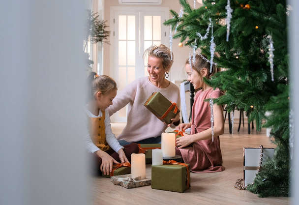 Alegre mamá y sus hijas lindas intercambian regalos de Navidad sentados en el suelo cerca del árbol de Navidad. - Foto, imagen