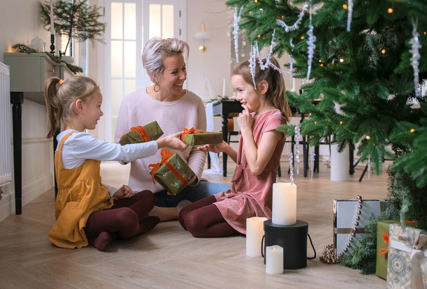 Vidám anya és aranyos lányai karácsonyi ajándékokat cserélnek a padlón ülve a karácsonyfa mellett.. - Fotó, kép