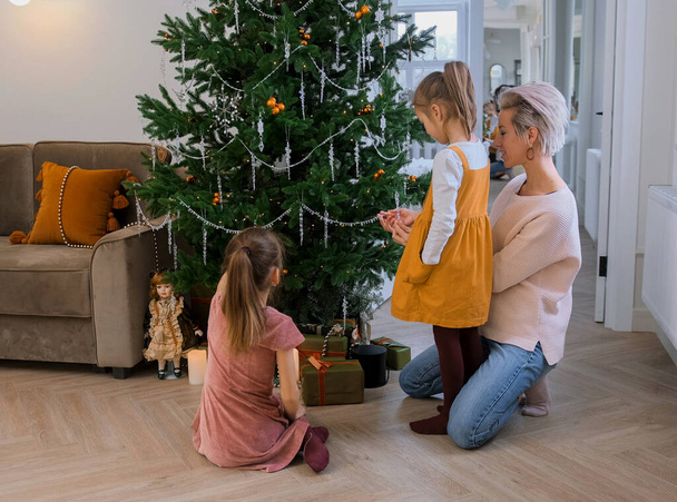 Η μαμά και οι κόρες διακοσμούν ένα χριστουγεννιάτικο δέντρο που κάθεται στο πάτωμα στο σαλόνι. - Φωτογραφία, εικόνα