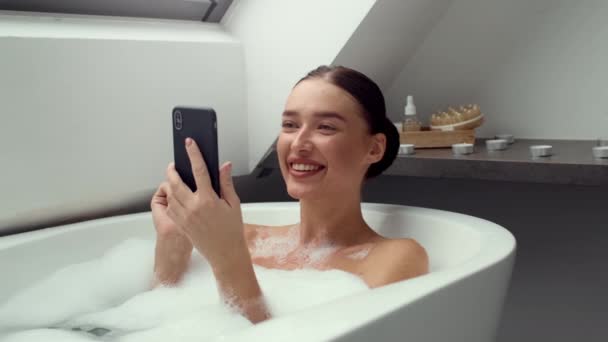 4K відеозапис, щаслива молода жінка, занурена в пінисту ванну, дивлячись на її смартфон, повільний рух. Сцена зображує суміш релаксації та сучасного цифрового зв'язку в моменти дозвілля. - Кадри, відео