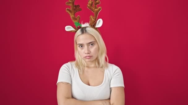 Young beautiful hispanic woman wearing reindeer christmas hat standing with serious expression and arms crossed gesture over isolated red background - Footage, Video