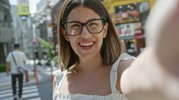 Stadsgezicht van tokyo fleurt als mooie Spaanse vrouw, versieren bril, vrolijk bezig met een leuke video gesprek. haar glimlachende, zelfverzekerde uitdrukking straalt geluk uit op de stedelijke straat. - Video