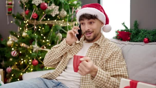 Молодой араб, разговаривающий по смартфону, пьет кофе, сидя дома у рождественской елки - Кадры, видео