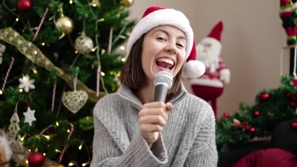 Νεαρή όμορφη ισπανίδα τραγουδίστρια που κάθεται στον καναπέ δίπλα στο χριστουγεννιάτικο δέντρο στο σπίτι - Πλάνα, βίντεο