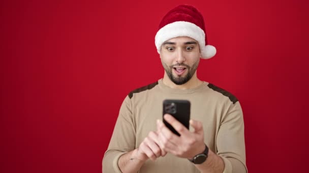 młody arabski człowiek noszący świąteczny kapelusz za pomocą smartfona na izolowanym czerwonym tle - Materiał filmowy, wideo