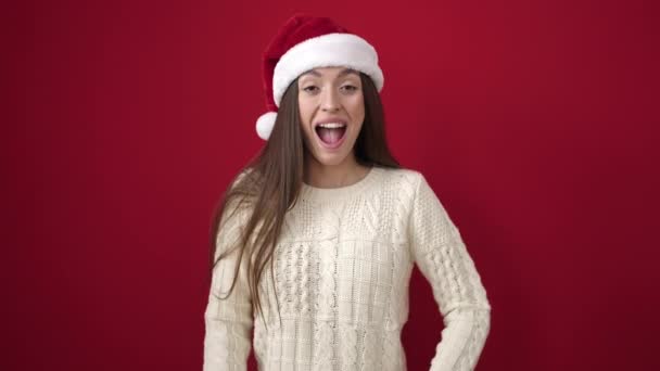 Junge schöne hispanische Frau lächelt selbstbewusst mit Weihnachtsmütze über isoliertem rotem Hintergrund - Filmmaterial, Video