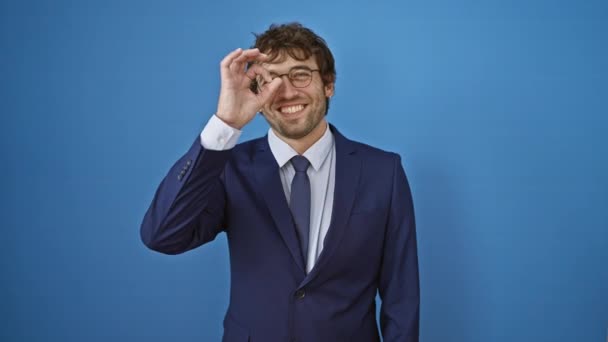 Veselý mladý muž v obleku bliká šťastný ok znamení mezi prsty, usmívá se na osamělém modrém pozadí, baví se s tímto humorným pohledem - Záběry, video