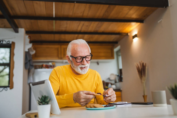 Een oudere man Kaukasische oude man gepensioneerde zit aan een tafel thuis zetten jam op een koekje voor te bereiden op het ontbijt genieten van snack ontspannen in zijn huis omgeving echte persoon kopiëren ruimte dagelijks routine - Foto, afbeelding