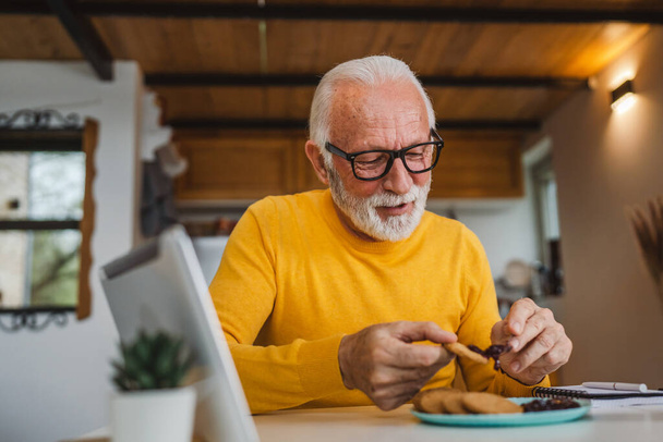 Een oudere man Kaukasische oude man gepensioneerde zit aan een tafel thuis zetten jam op een koekje voor te bereiden op het ontbijt genieten van snack ontspannen in zijn huis omgeving echte persoon kopiëren ruimte dagelijks routine - Foto, afbeelding