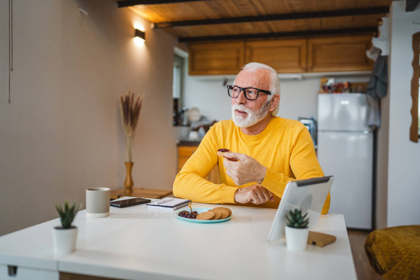 Ένας ηλικιωμένος άνδρας καυκάσιος γέρος συνταξιούχος κάθεται σε ένα τραπέζι στο σπίτι βάζοντας μαρμελάδα σε ένα μπισκότο που ετοιμάζεται να φάει πρωινό απολαύστε ένα σνακ χαλαρό στο περιβάλλον του σπιτιού του πραγματικό πρόσωπο αντιγραφή χώρου καθημερινή ρουτίνα - Φωτογραφία, εικόνα