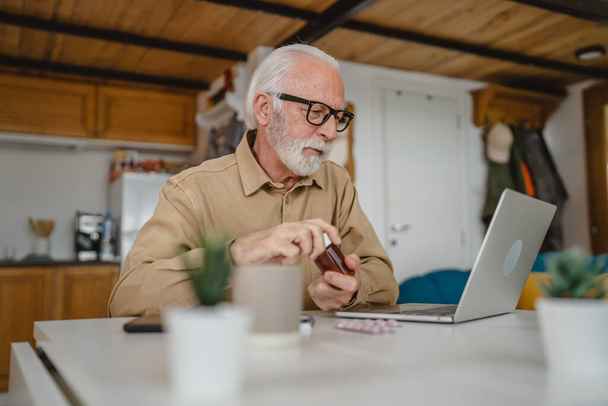 Jeden senior mężczyzna kaukaski mężczyzna dziadek siedzieć w domu trzymać paczkę tabletek przeciwbólowych lub witaminy suplement czytać etykieta gotowy do podjęcia leków w pobliżu laptop zamówić online przestrzeń kopia - Zdjęcie, obraz